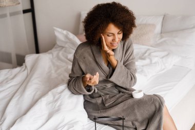 Kolajen kaldırma kremi olan Afrikalı bir kız yatak odasında sabah güzelliğini rutin hale getiriyor. Bornozlu Afrikalı Amerikalı kadın yüzüne temizleme ve gençleştirme kremi sürüyor.