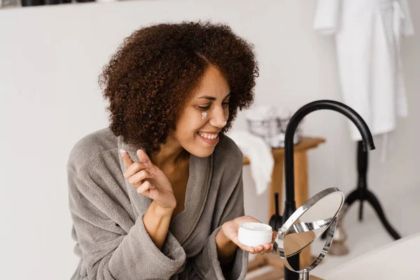 African girl applying face moisturizer cream to protect skin from dryness in the bath. African american woman in bathrobe with facial moisturizing cream doing morning beauty routine