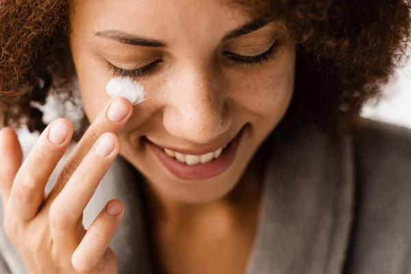 African girl applying face moisturizer cream close-up to protect skin from dryness in bath. African american woman in bathrobe with facial moisturizing cream doing morning beauty routine