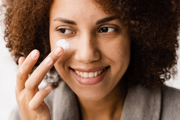 stock image African girl applying face moisturizer cream close-up to protect skin from dryness in bath. African american woman in bathrobe with facial moisturizing cream doing morning beauty routine