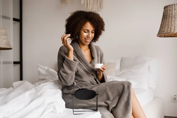 African american woman in bathrobe with facial moisturizing cream doing morning beauty routine in bedroom. African girl applying face moisturizer cream to protect skin from dryness