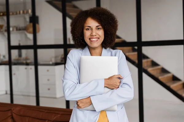 Candid african american girl with laptop standing in office, smiles and rejoices at successful work in IT company. Cheerful african young woman programmer is standing with laptop and smiling