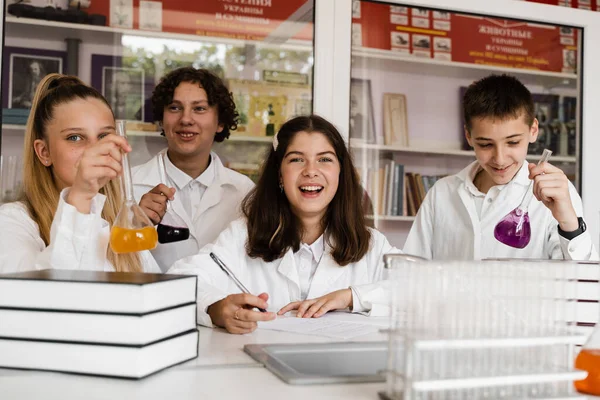 Schoolchildren studying at chemistry lesson in classrom. Pupils writing in notebook, holding flasks with liquid for experiments and have fun together. School education