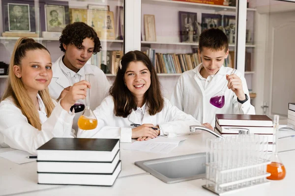Schoolchildren studying at chemistry lesson in classrom. Pupils writing in notebook, holding flasks with liquid for experiments and have fun together. School education