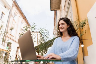 Neşeli genç bayan programcı dizüstü bilgisayarla uzaktan çalışıyor ve dışarıda kafede son teslim tarihine yetişmeye çalışıyor. Bilgisayarlı samimi kız gülümsüyor ve IT şirketinde başarılı çalışmaktan memnun.