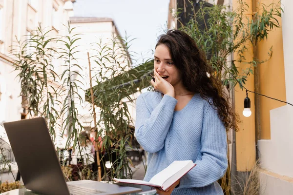 Cheerful surprised woman smiling and looking at amount of income earnings in online casino and rejoicing outside. Shocked girl is winning bet in online casino and sitting with laptop in cafe outdoor