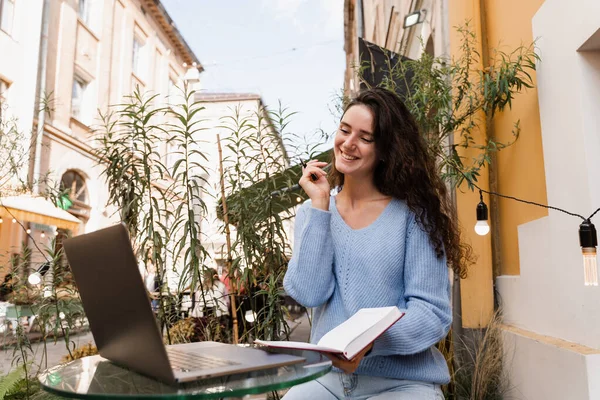 Shocked girl is winning bet in online casino and sitting with laptop in cafe outdoor. Cheerful surprised woman smiling and looking at amount of income earnings in online casino and rejoicing outside