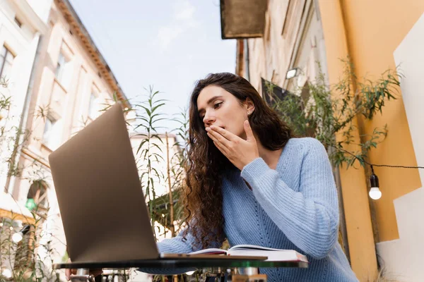 Shocked girl is winning bet in online casino and sitting with laptop in cafe outdoor. Cheerful surprised woman smiling and looking at amount of income earnings in online casino and rejoicing outside