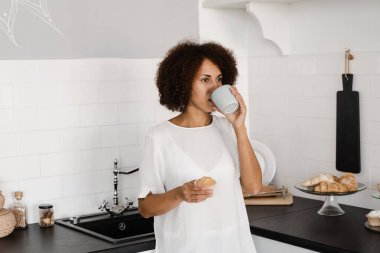 Beyaz mutfakta kahve ve kekle çekici bir Afrikalı kadın. Afro-Amerikan kızıyla sıcak bir sabah. Evdeki mutfakta.