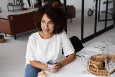 Afrikalı genç bir kadın kahvaltı yapıyor ve tüm gün boyunca enerji takviyesi alıyor. Afro-Amerikalı kız kahve içiyor ve yemek masasında kek tadıyor. Rahat sabah yaşam tarzı