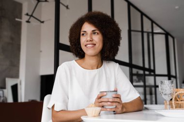 Afrikalı genç bir kadın kahvaltı yapıyor ve tüm gün boyunca enerji takviyesi alıyor. Afro-Amerikalı kız kahve içiyor ve yemek masasında kek tadıyor. Rahat sabah yaşam tarzı