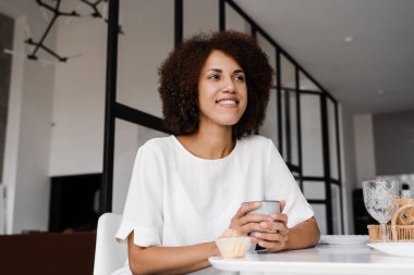 Afrikalı genç bir kadın kahvaltı yapıyor ve tüm gün boyunca enerji takviyesi alıyor. Afro-Amerikalı kız kahve içiyor ve yemek masasında kek tadıyor. Rahat sabah yaşam tarzı