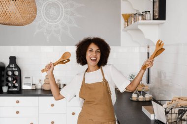Afro-Amerikalı aşçı kız spatulayla yemek pişirmek ve eğlenmek için dans ediyor. Önlüklü mutlu Afrikalı ev kadını kahvaltı hazırlamadan önce mutfakta şarkı söyleyip dans ediyor.