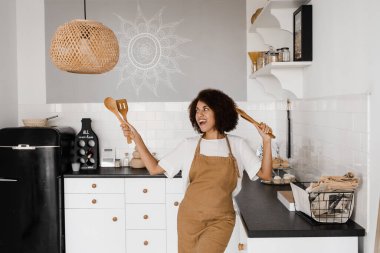 Afro-Amerikalı aşçı kız spatulayla yemek pişirmek ve eğlenmek için dans ediyor. Önlüklü mutlu Afrikalı ev kadını kahvaltı hazırlamadan önce mutfakta şarkı söyleyip dans ediyor.