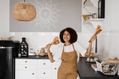 Afro-Amerikalı aşçı kız spatulayla yemek pişirmek ve eğlenmek için dans ediyor. Önlüklü mutlu Afrikalı ev kadını kahvaltı hazırlamadan önce mutfakta şarkı söyleyip dans ediyor.