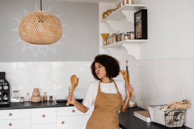 Önlüklü mutlu Afrikalı ev hanımı kahvaltı hazırlamadan önce mutfakta şarkı söyleyip dans ediyor. Afro-Amerikalı aşçı kız spatulalarla dans ediyor ve yemek pişiriyor.
