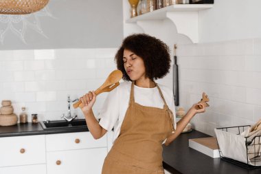Önlüklü Afrikalı Amerikalı ev hanımı mutfakta mikrofon pişirmek için spatulalarla şarkı söylüyor. Neşeli Afrikalı genç kadın mutfak aletleriyle eğleniyor.