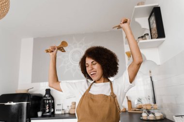 Afrikalı Amerikalı kadın mutfakta dans ediyor ve ellerini kaldırıyor. Önlüklü Afrikalı bir kız beyaz mutfakta dans ediyor. Yemek kursları ve yemek atölyeleri için reklam.