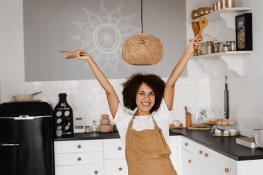 Afrikalı Amerikalı kadın mutfakta dans ediyor ve ellerini kaldırıyor. Önlüklü Afrikalı bir kız beyaz mutfakta dans ediyor. Yemek kursları ve yemek atölyeleri için reklam.