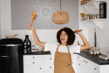 Önlüklü Afrikalı bir kız beyaz mutfakta dans ediyor. Yemek kursları ve yemek atölyeleri için reklam. Afrikalı Amerikalı kadın mutfakta dans ediyor ve ellerini kaldırıyor.
