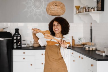 Önlüklü Afrikalı bir kız beyaz mutfakta dans ediyor. Yemek kursları ve yemek atölyeleri için reklam. Afrikalı Amerikalı kadın mutfakta dans ediyor ve ellerini kaldırıyor.