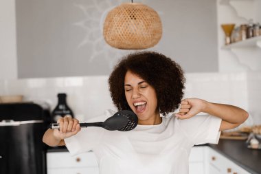 Neşeli Afrikalı genç bir kadın mutfak aletleriyle eğleniyor. Önlüklü Afrikalı Amerikalı ev hanımı mutfakta mikrofon pişirmek için spatula kullanıyor.