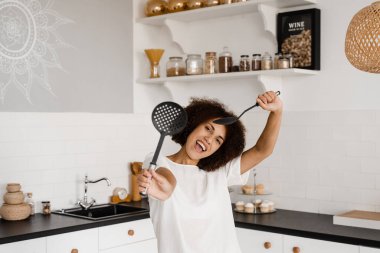 Önlüklü Afrikalı ev hanımı mutfakta yemek pişirmek için spatulalarla poz veriyor. Afro-Amerikalı aşçı kız mutfakta eğleniyor. Yemek kursları ve yemek atölyeleri için reklam.