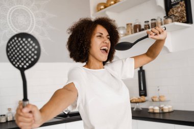 Neşeli Afrikalı genç bir kadın mutfak aletleriyle eğleniyor. Önlüklü Afrikalı Amerikalı ev hanımı mutfakta mikrofon pişirmek için spatula kullanıyor.