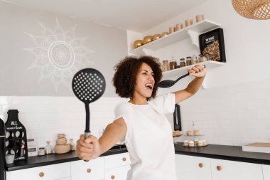 Neşeli Afrikalı genç bir kadın mutfak aletleriyle eğleniyor. Önlüklü Afrikalı Amerikalı ev hanımı mutfakta mikrofon pişirmek için spatula kullanıyor.