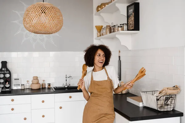 stock image Happy african housewife in apron singing and dancing on the kitchen before cooking breakfast. African american cook girl dancing with spatulas for cooking and having fun