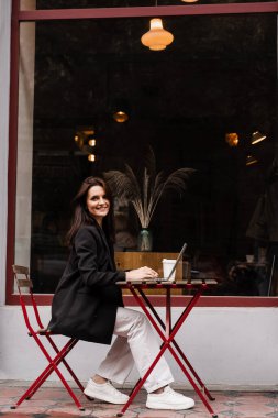 Laptoplu ve kahveli çekici kız kafenin dışında oturuyor. Bilgisayarlı genç iş kadını dışarıda çalışıyor ve kahve içiyor.