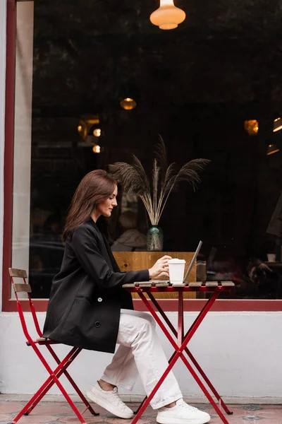 Laptoplu ve kahveli çekici kız kafenin dışında oturuyor. Bilgisayarlı genç iş kadını dışarıda çalışıyor ve kahve içiyor.