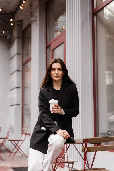 stock image Confident young business woman with cup of coffee is posing outside of cafe on terrace. Cheerful girl is weared casual business style is posing near cafe