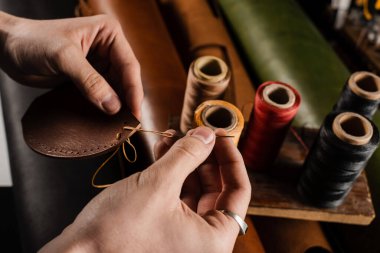 Craftsman sews genuine leather using needle and thread for creation natural leather products. Equipment for genuine leather production in workshop. Process of stitching genuine leather