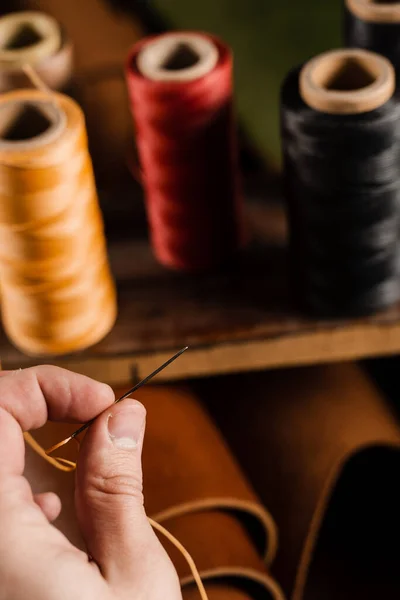 Craftsman is holding needle with thread on roll of brown genuine leather background in craftsman workshop. Handmade genuine leather production in the factory