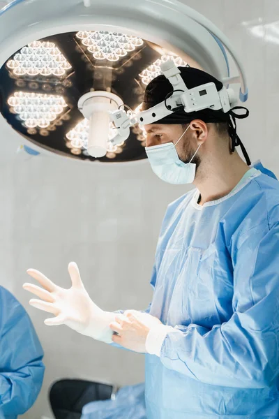 Male surgeon puts on surgical gloves and disinfects before operation. Surgeon with headlight is preparing for surgery in medical clinic