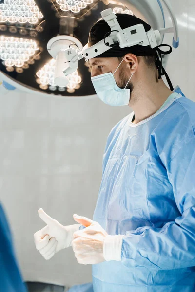 Surgeon with headlight is preparing for surgery in medical clinic. Male surgeon puts on surgical gloves and disinfects before operation