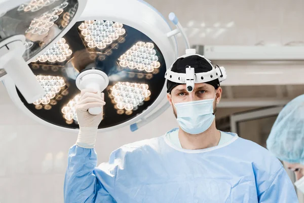 Male surgeon is holding light lamp in the operating room and directs it to the operating field. Surgeon with headlight is preparing for surgery in medical clinic