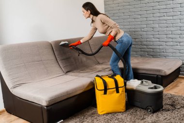 Cleaner girl is cleaning couch with extraction machine for dry clean upholstered furniture. Housekeeper is extracting dirt from upholstered sofa using dry cleaning extractor machine