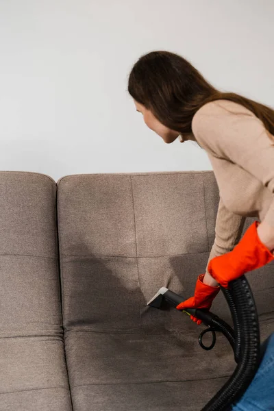 Cleaner girl is cleaning couch with extraction machine for dry clean upholstered furniture. Housekeeper is extracting dirt from upholstered sofa using dry cleaning extractor machine