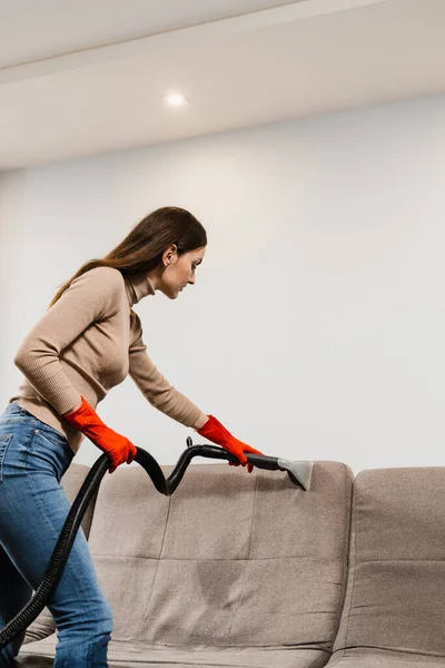 Cleaner girl is cleaning couch with extraction machine for dry clean upholstered furniture. Housekeeper is extracting dirt from upholstered sofa using dry cleaning extractor machine