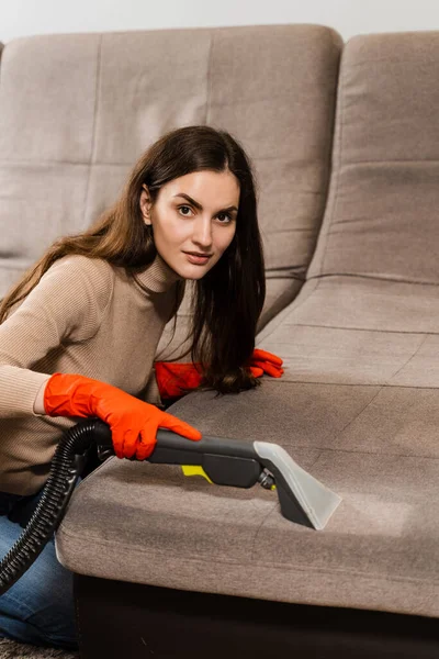 Cleaner girl is cleaning couch with extractor machine for dry clean upholstered furniture. Housekeeper is extracting dirt from upholstered sofa using dry cleaning extractor machine