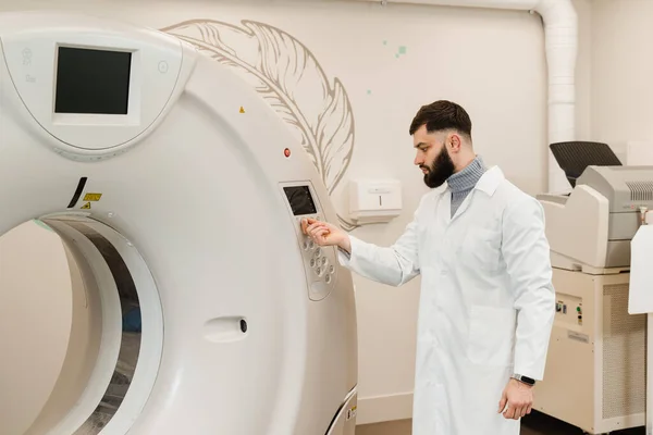 Man doctor adjusts CT computed tomography scanner in medical clinic. Handsome bearded radiologist in medical robe with computed tomograph for obtain detailed internal images of the body of patient