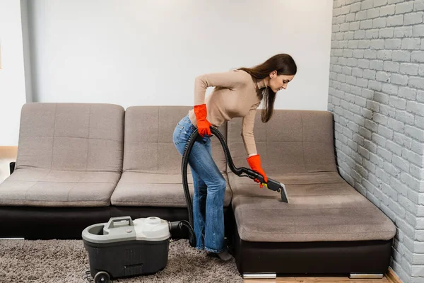 Cleaner girl is cleaning couch with extractor dry cleaning machine at clients home. Housewife dry cleaning of upholstered furniture and removing dust, stains and dirt using extractor machine