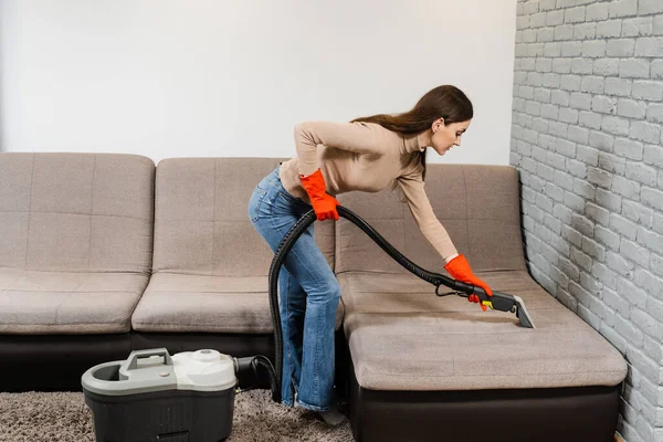 Housewife dry cleaning of upholstered furniture and removing dust, stains and dirt using extractor machine. Cleaner girl is cleaning couch with extractor dry cleaning machine at clients home
