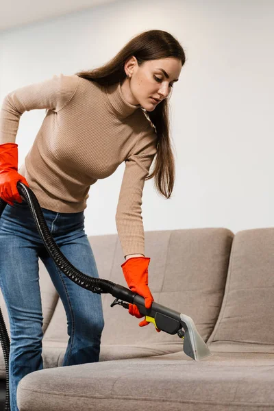 Cleaner girl is cleaning couch with extractor machine for dry clean upholstered furniture. Housekeeper is extracting dirt from upholstered sofa using dry cleaning extractor machine