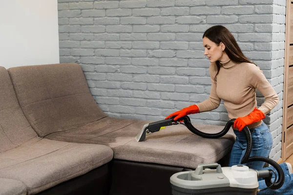 Cleaner girl is cleaning couch with extractor dry cleaning machine at clients home. Housewife dry cleaning of upholstered furniture and removing dust, stains and dirt using extractor machine