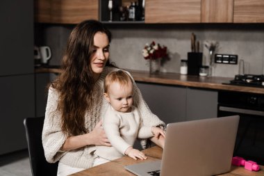 Mom and child daughter are watching cartoons on laptop at home in the kitchen. Motherhood. Family of mother and daughter toddler spending time together and watching videos online