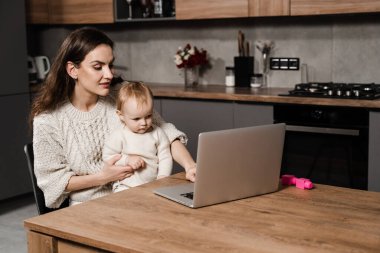 Family of mother and daughter toddler spending time together and watching videos online. Motherhood. Mom and child daughter are watching cartoons on laptop at home in the kitchen