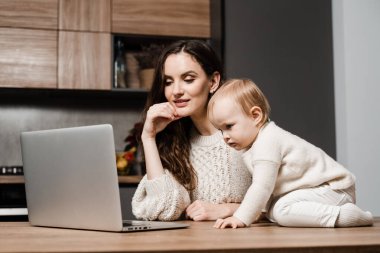 Mom and child daughter are watching cartoons on laptop at home in the kitchen. Motherhood. Family of mother and daughter toddler spending time together and watching videos online